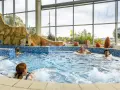 Ludzie relaksują się w basenie termalnym z hydromasażem, otoczonym skalnymi formacjami i dużymi oknami.