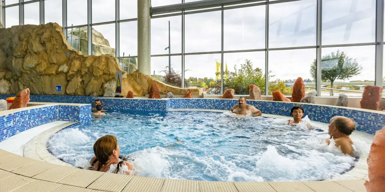 Ludzie relaksują się w basenie termalnym z hydromasażem, otoczonym skalnymi formacjami i dużymi oknami.