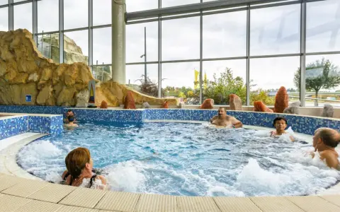 Ludzie relaksują się w basenie termalnym z hydromasażem, otoczonym skalnymi formacjami i dużymi oknami.