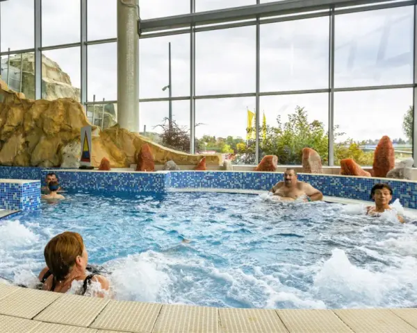 Ludzie relaksują się w basenie termalnym z hydromasażem, otoczonym skalnymi formacjami i dużymi oknami.